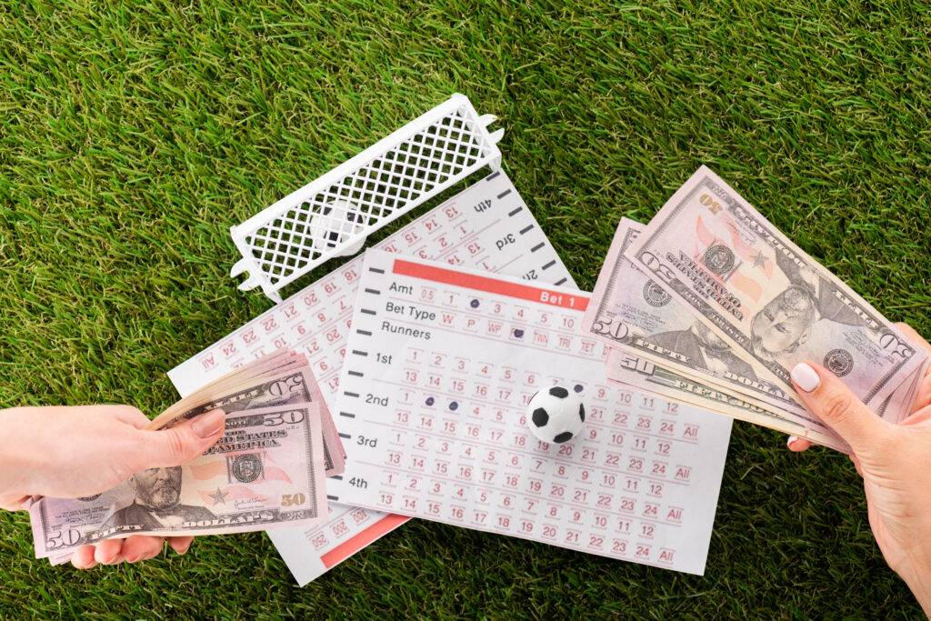 Two hands holding American dollar in front of two betting slips. On one of the betting slips is a toy football ball, and beside the other slide is a small plastic toy football goal with a toy football ball in it. This is all on top of a green grass background.