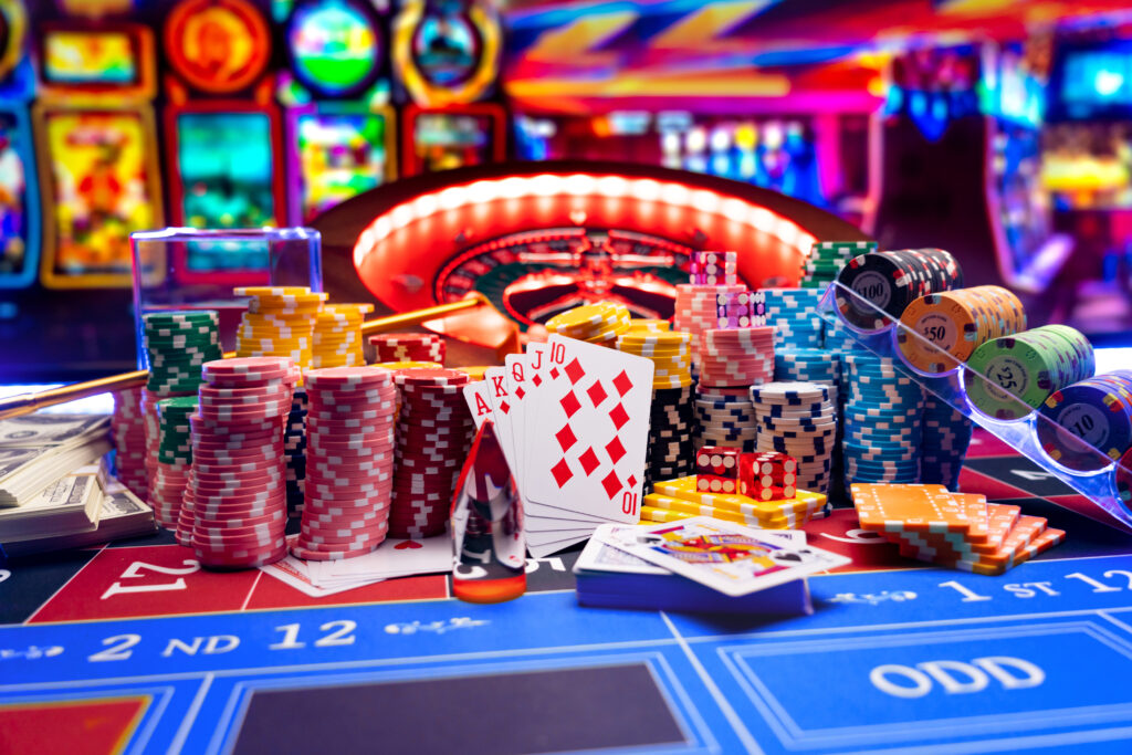 Collection of playing cards, casino chips, dollars and dice in front of an angled roulette wheel on a roulette table. In the background are slot machines, which are blurred.